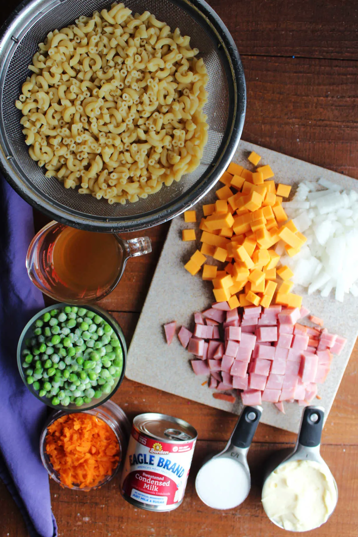 Ingredients including cooked macaroni noodles, peas, vinegar, shredded carrots, sweetened condensed milk, sugar, mayonnaise, ham, cubed cheddar cheese, and diced onion ready to be made into sweet pasta salad. 