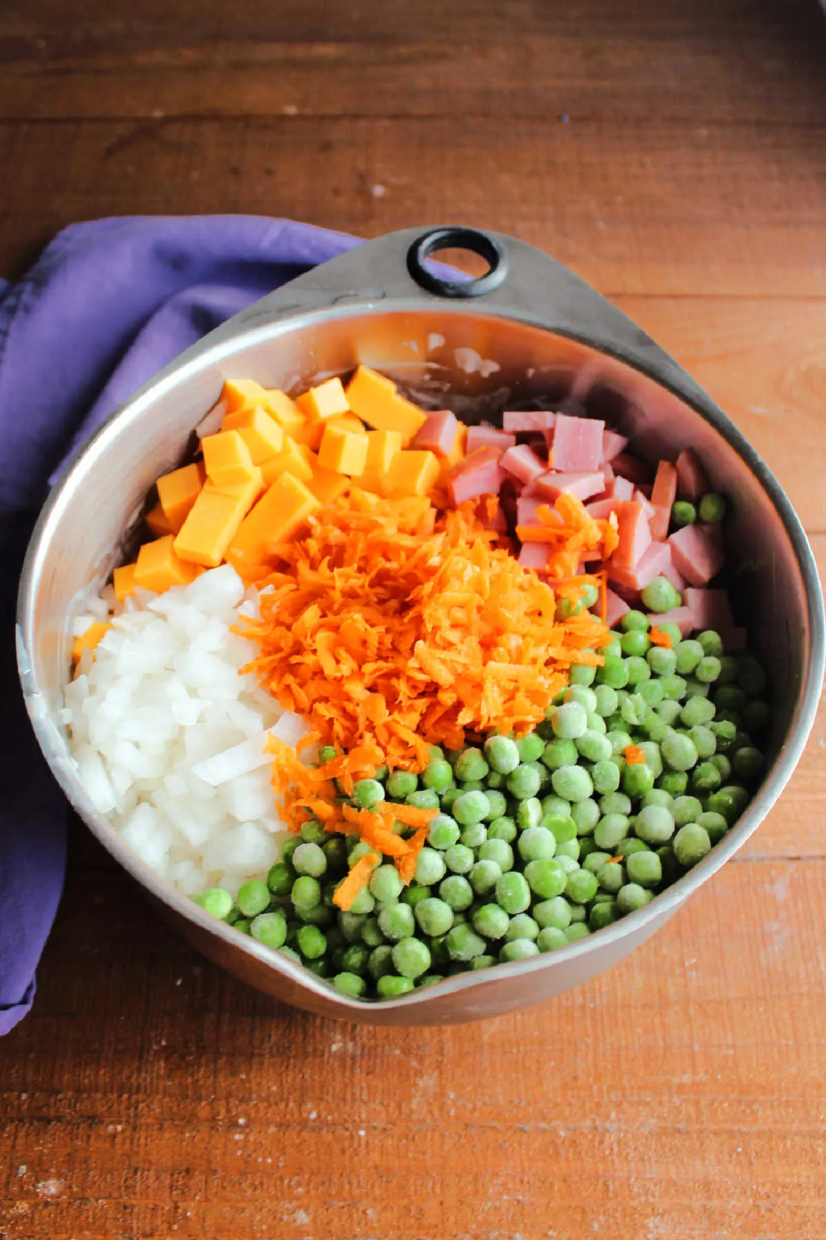 Frozen peas, shredded carrots, cubed ham and cheddar, and diced onion on top of pasta and dressing ready to be mixed in.