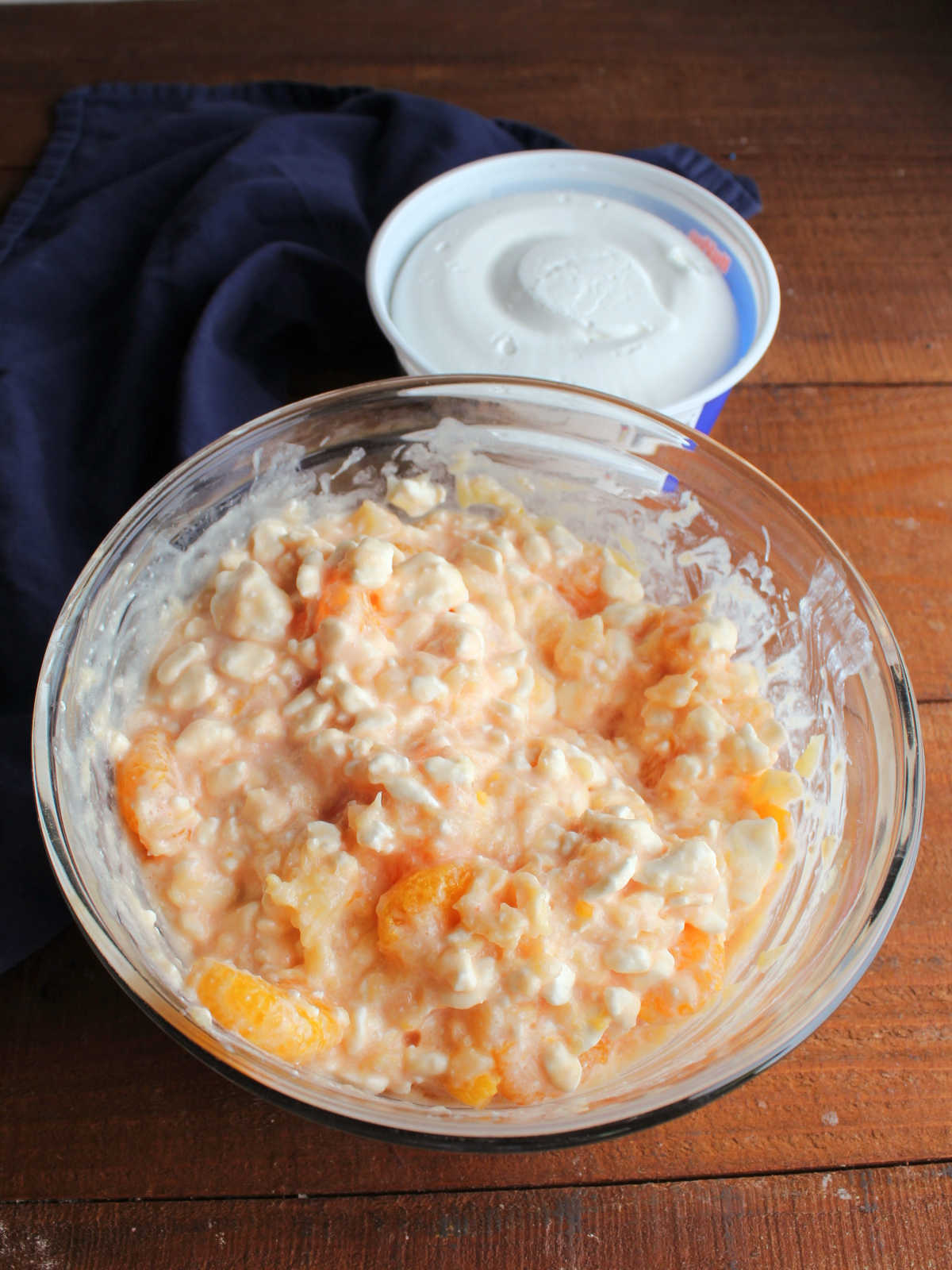 Drained cans of crushed pineapple and mandarin oranges stirred into the orange cottage cheese with a tub of whipped topping in the background. 