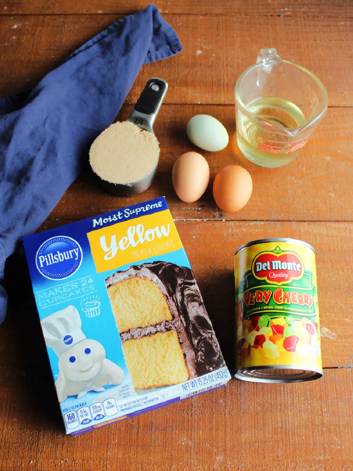 Ingredients including yellow cake mix, eggs, oil, can of fruit cocktail, and brown sugar ready to be made into fruit cocktail cake.