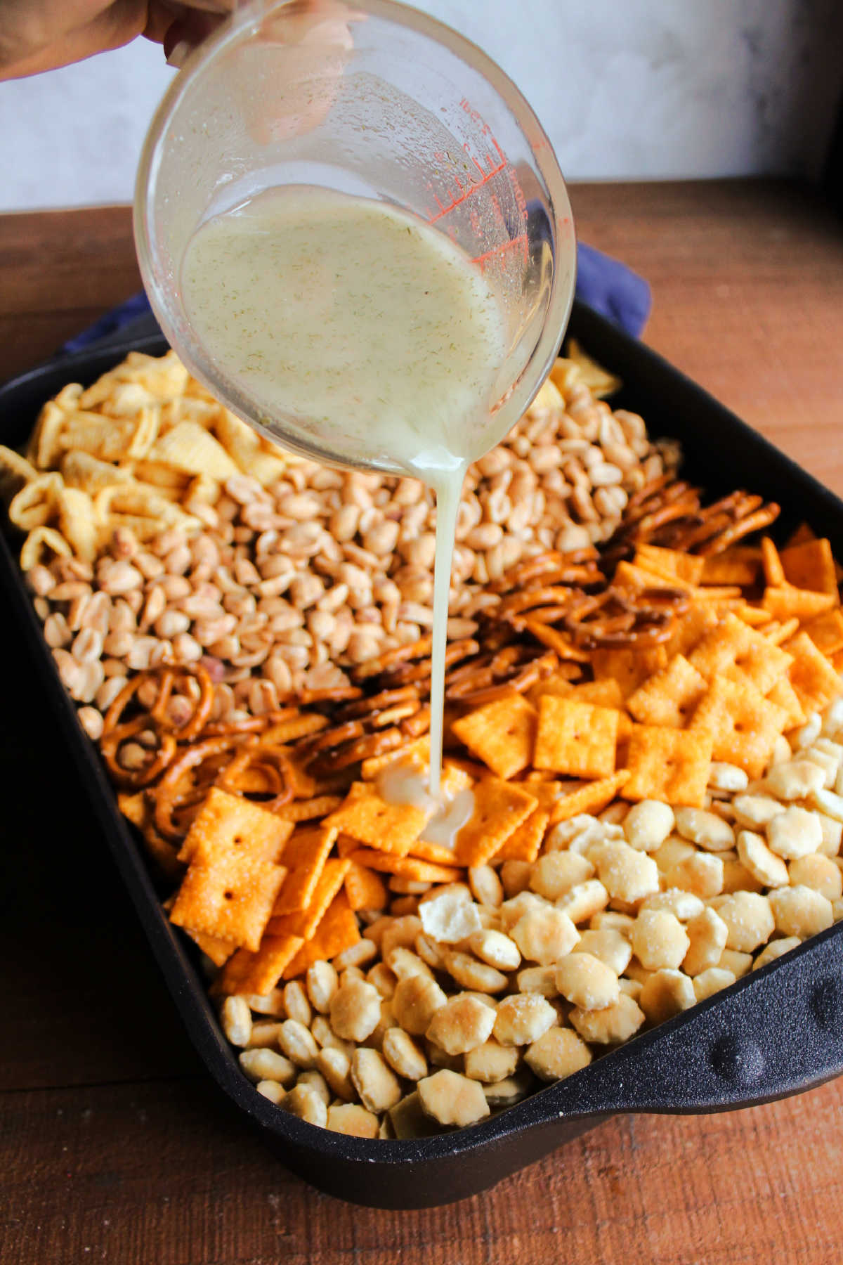 Pouring oil mixed with ranch seasoning over peanuts and crackers in roasting pan.