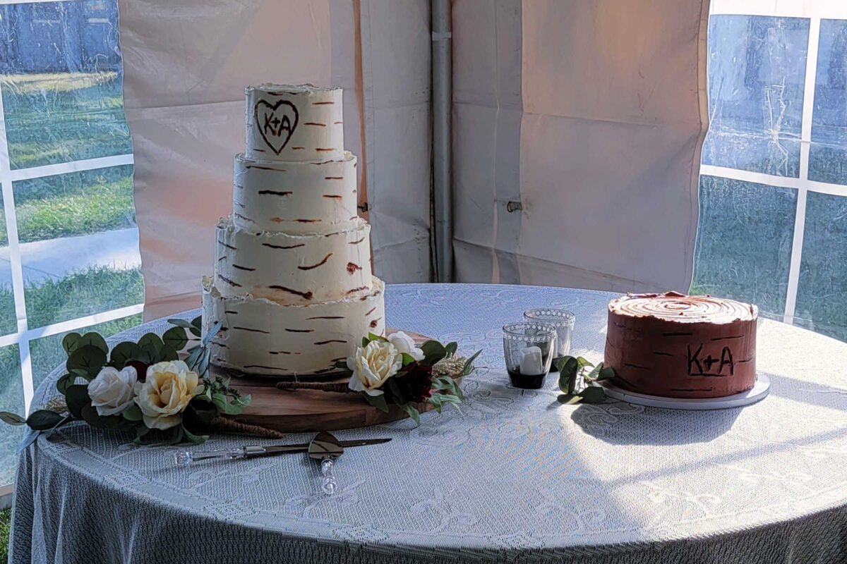 Wedding cake table with 4 tier vanilla cake with birch tree bark looking finish and carved in initials next to a 10 inch chocolate groom's cake with similar birch look and carved initials. 