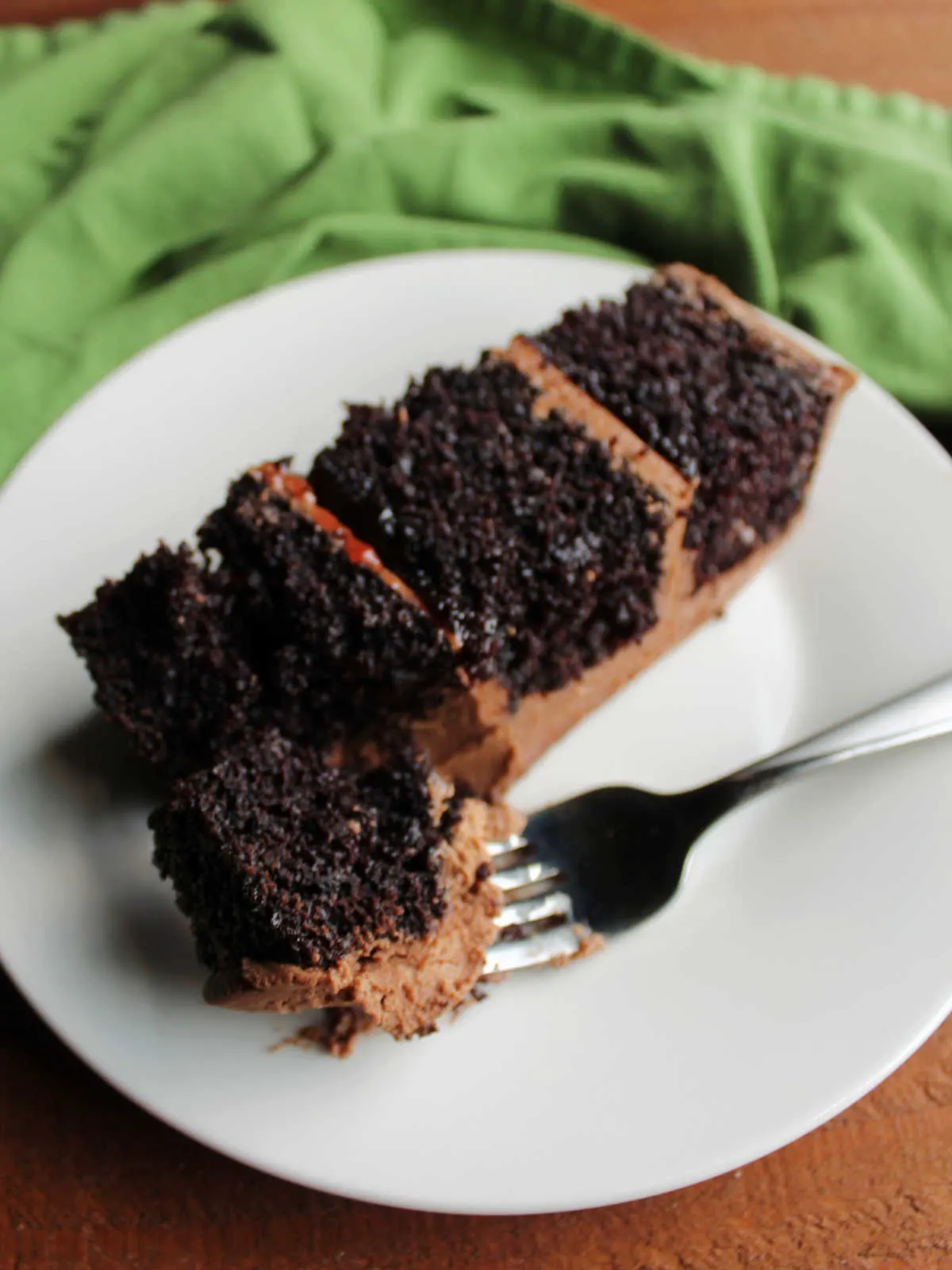 Bite of rich chocolate cake on fork, ready to eat with remaining slice on plate showing dark chocolate cake, plenty of chocolate buttercream, and some raspberry filling.