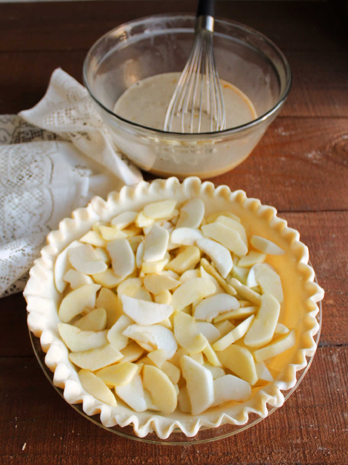 Pie crust filled with apple slices with bowl of custard mixture in the background.
