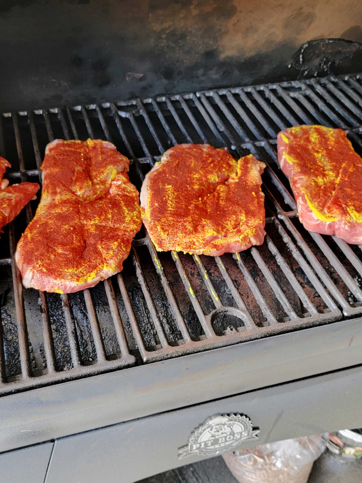 Pork steaks coated in mustard and dry rub on the smoker grates, ready to start cooking.