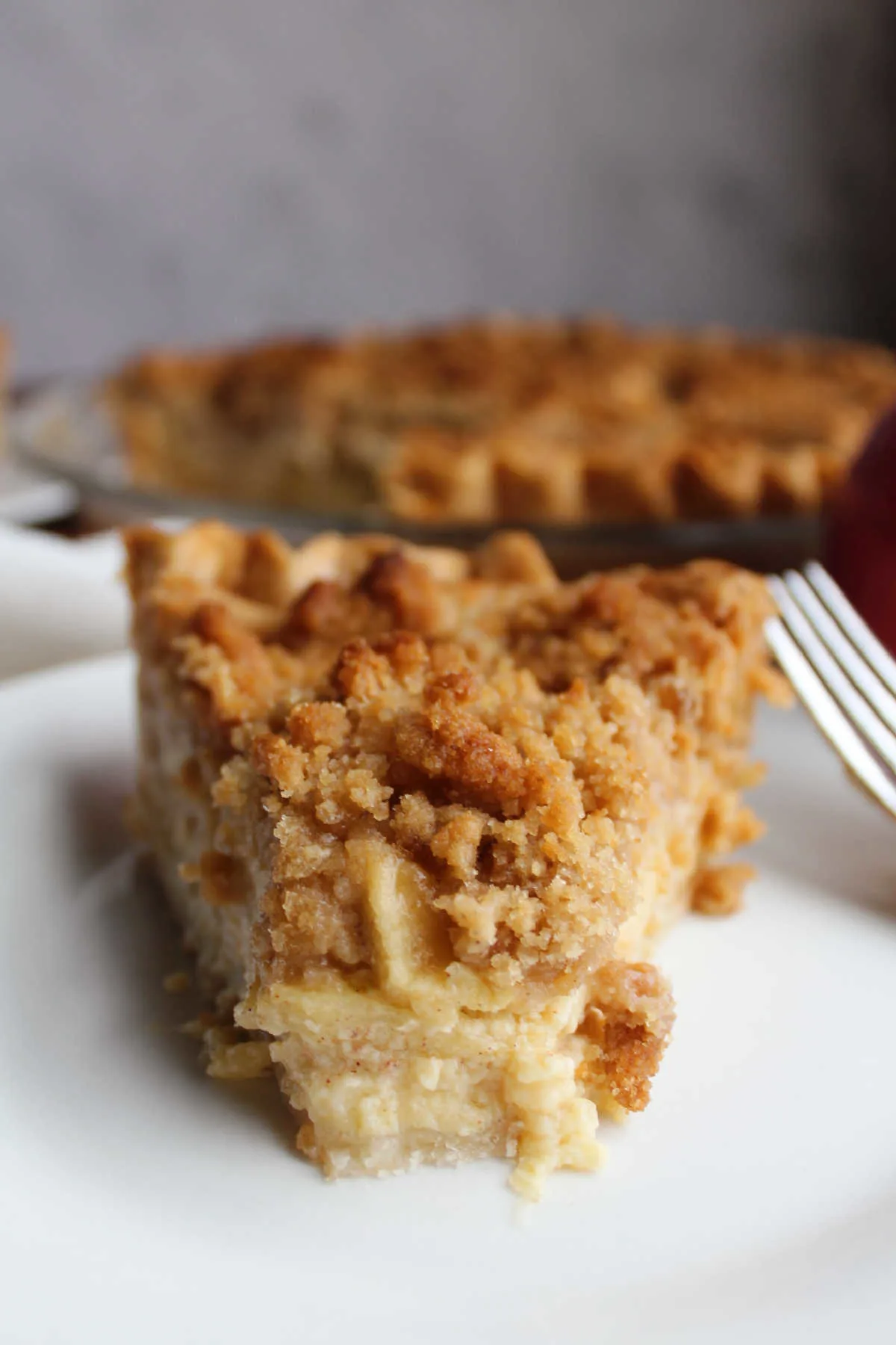 Slice of apple custard pie on plate with bite missing from tip showing creamy custard surrounding the apples and a golden streusel on top.