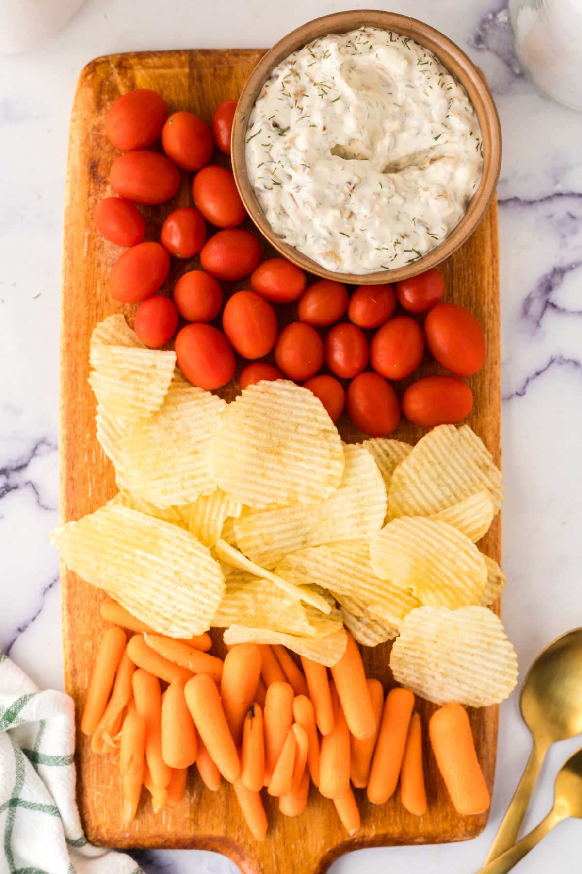Snack board with bowl of caramelized onion dip, cherry tomatoes, potato chips, and baby carrots.