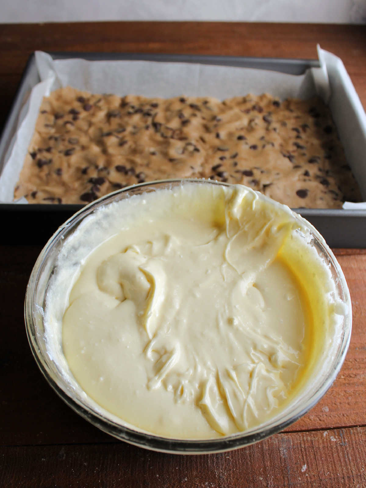 Mixing bowl with smooth cream cheese mixture in front of pan with chocolate chip cookie dough crust. 