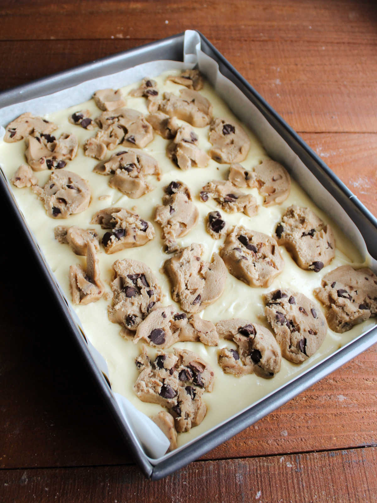 Chocolate chip cookie dough distributed over cheesecake mixture in pan, ready to bake.