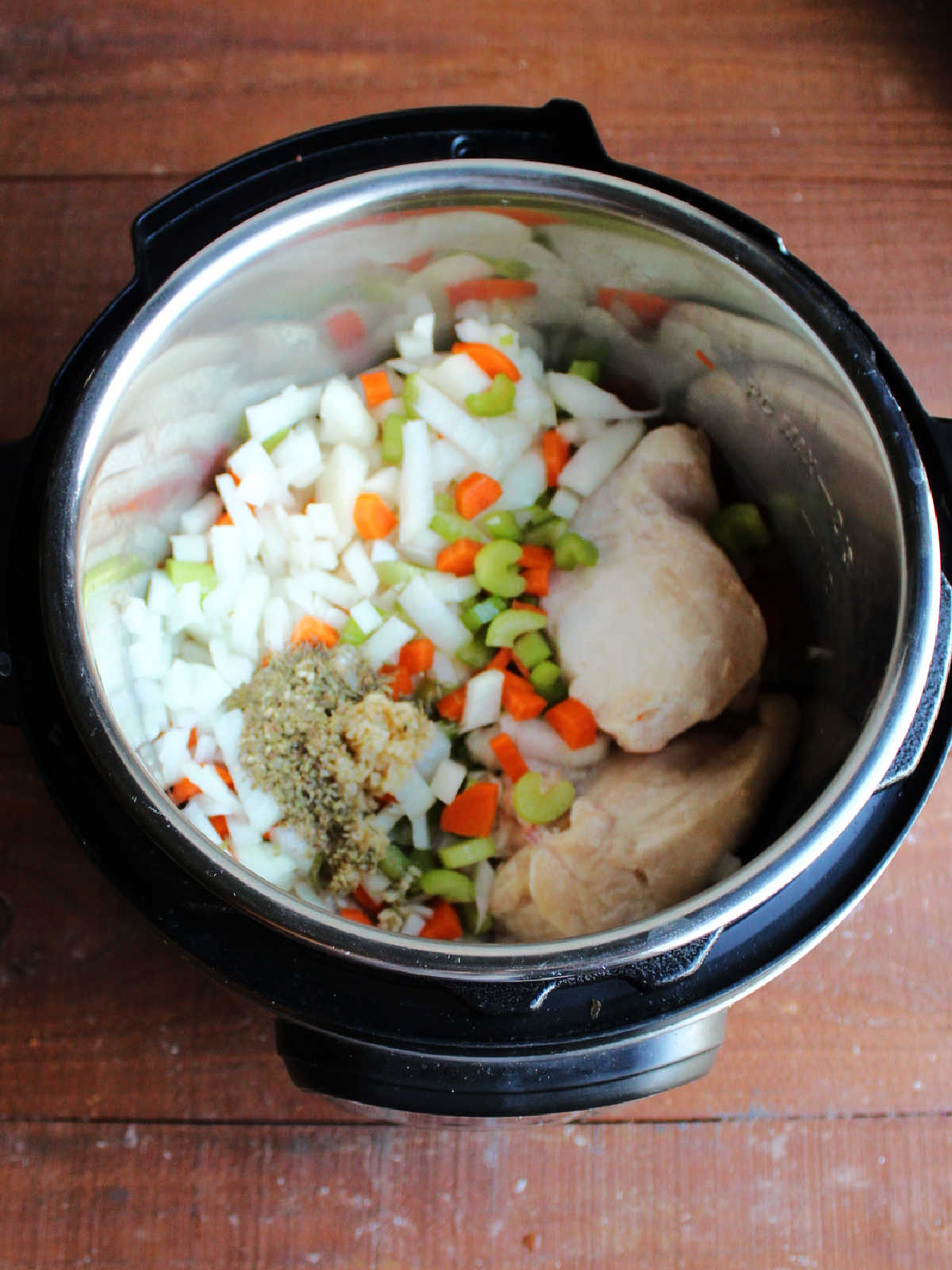 Instant pot with frozen chicken breasts, diced onion, sliced carrots and celery, herbs, and garlic inside ready for to be cooked.