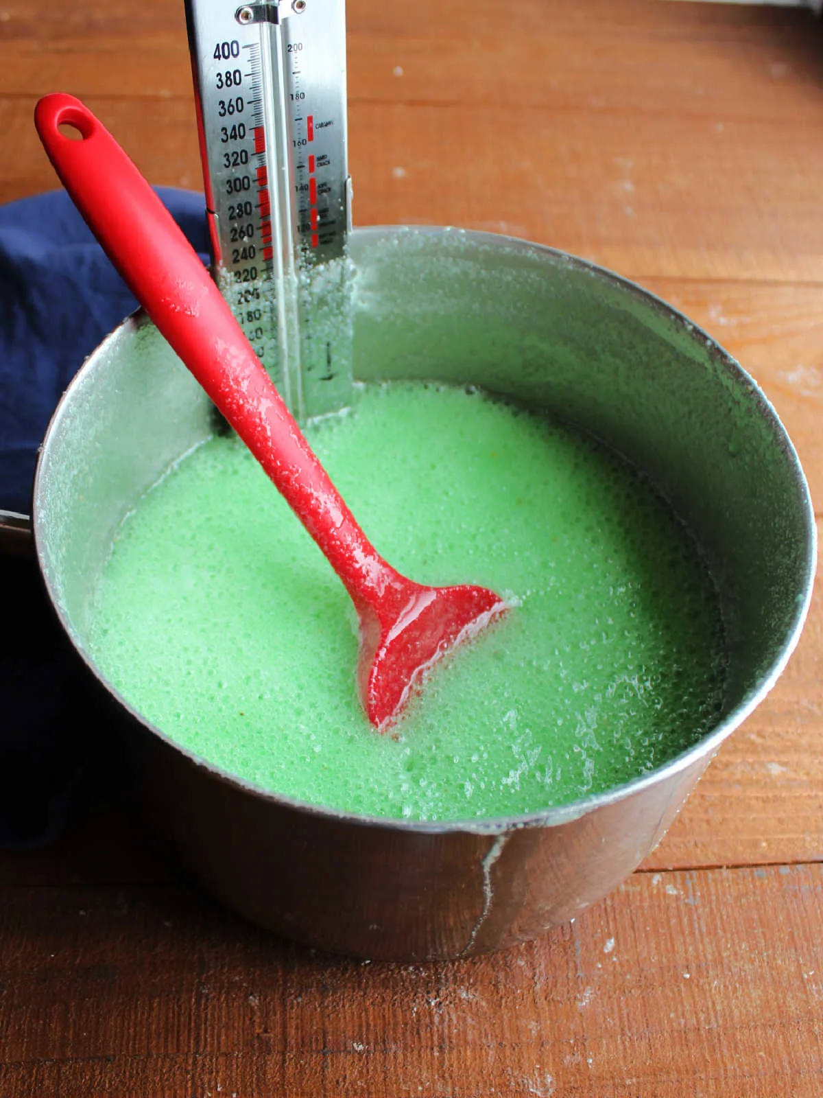 Saucepan with hot and foamy green jello mixture and candy thermometer inside.