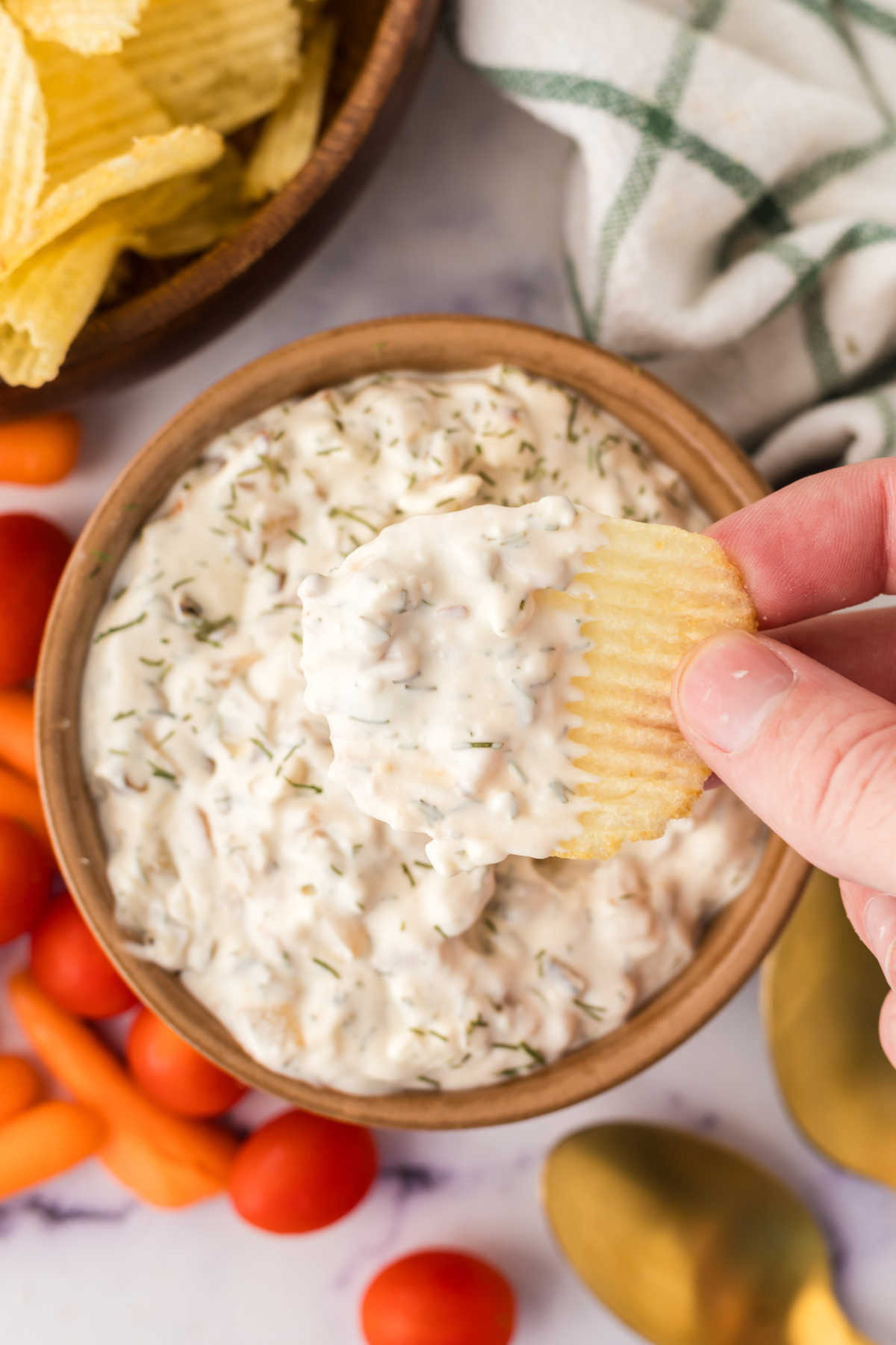 Hand holding ruffled potato chip with creamy onion dip on it, ready to eat.