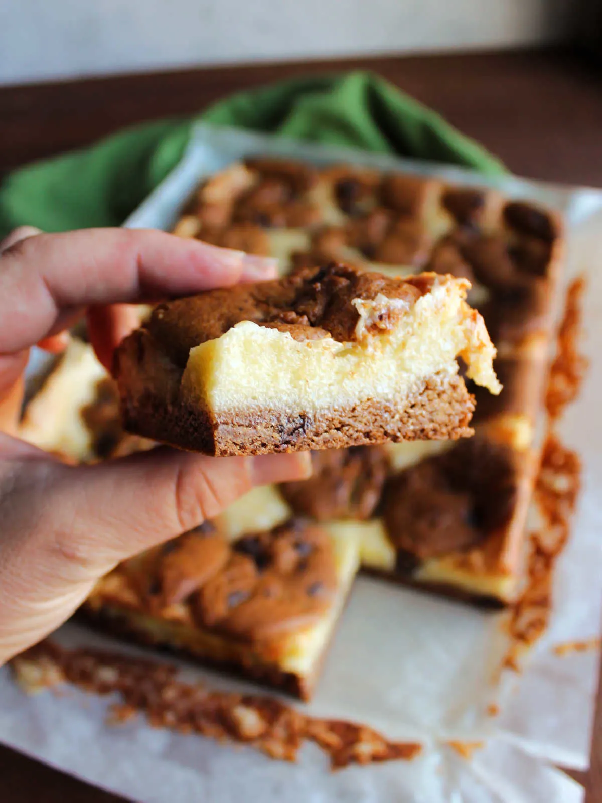 Hand holding cheesecake bar showing chocolate chip cookies surrounding smooth cream cheese filling.