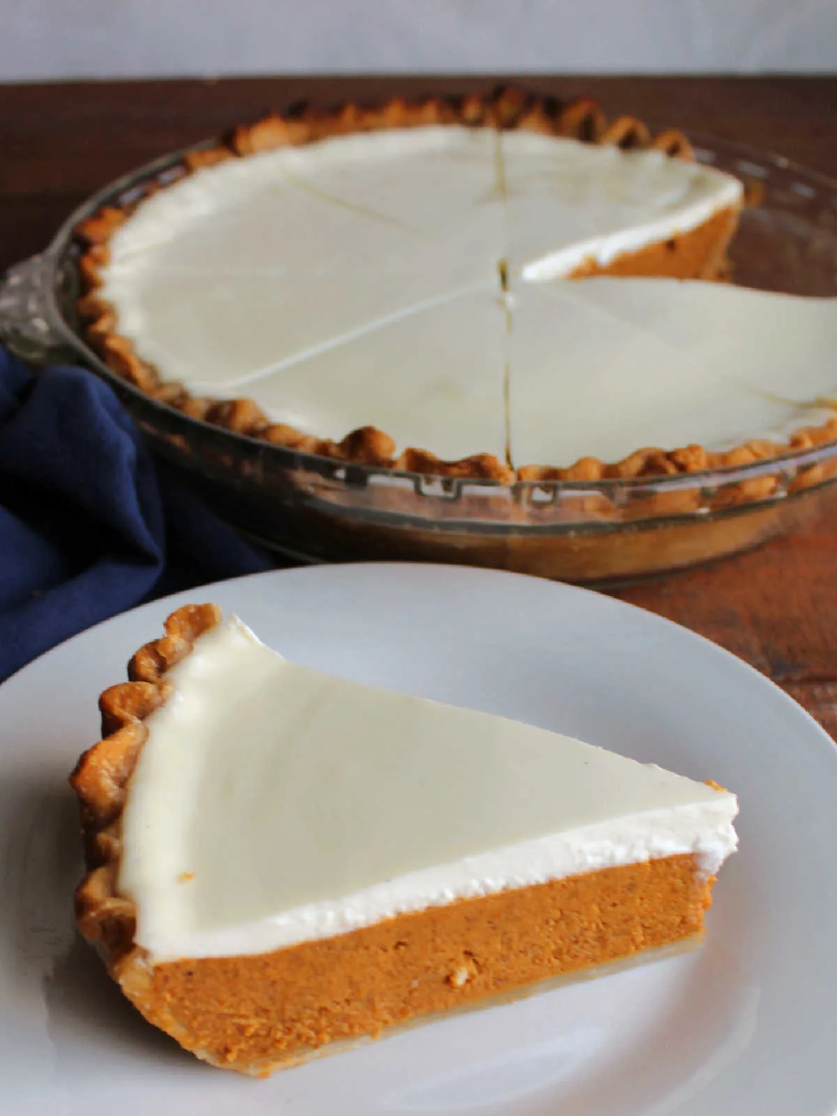 Piece of layered pumpkin pie with sour cream topping served on white plate with remaining pie in background. 