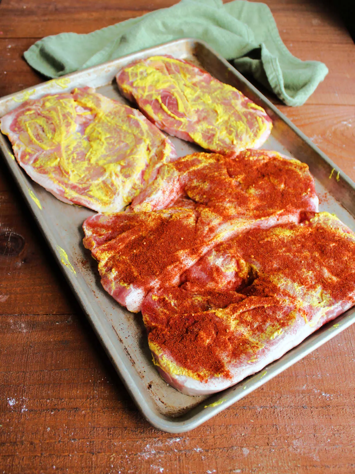 Pan of pork steaks coated in yellow mustard, two that have bbq rub on them and two that still need the rub applied.