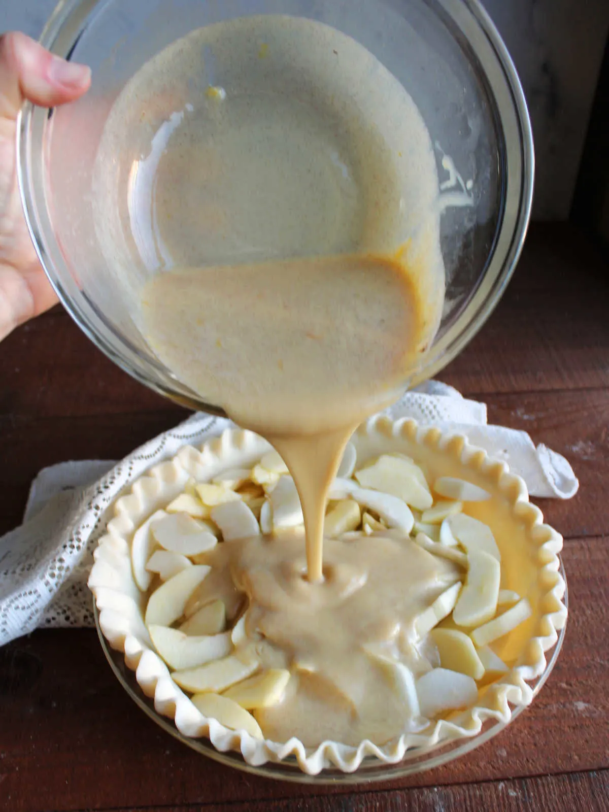 Pouring condensed milk custard over apple slices in pie plate.