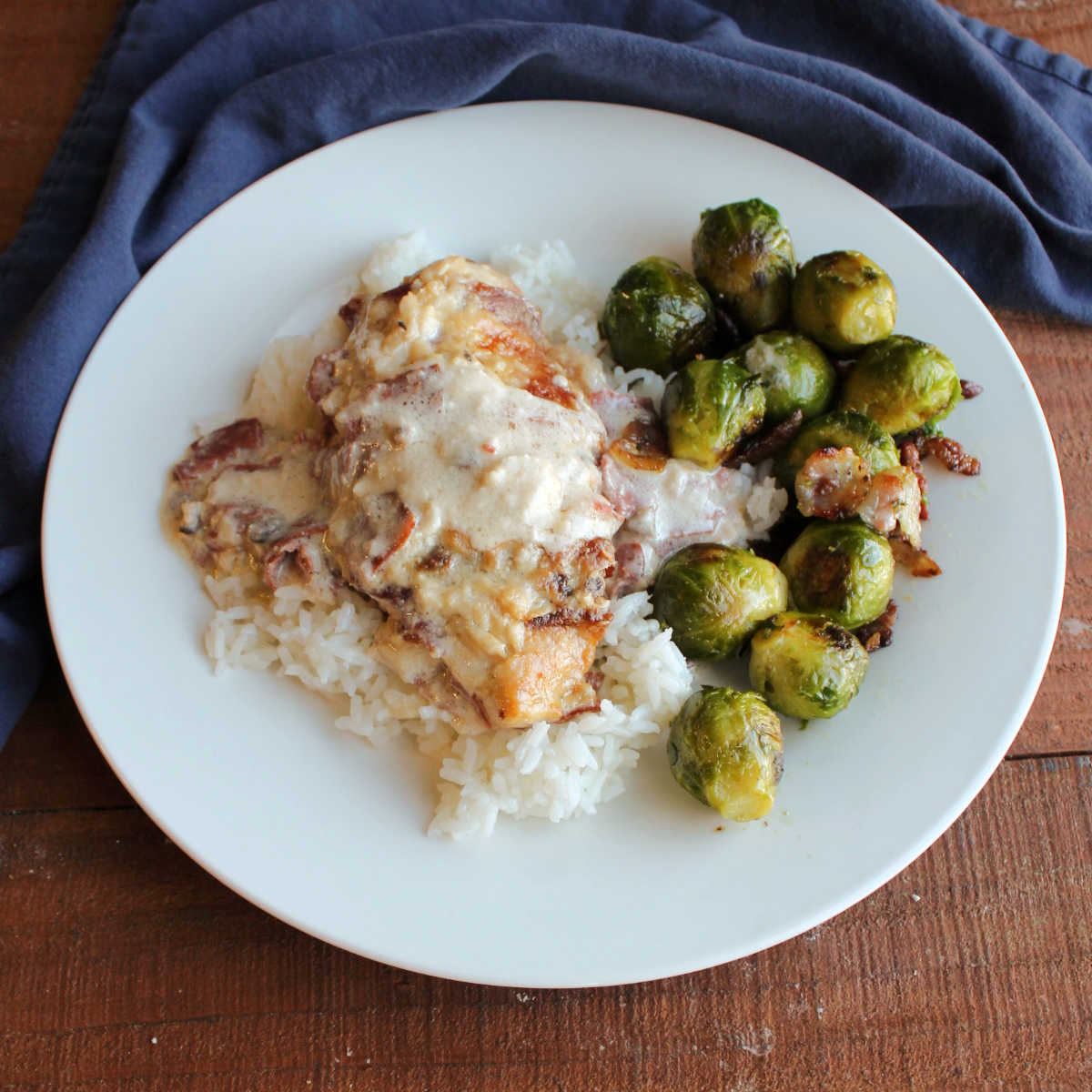 Dinner plate with bacon wrapped chicken breast in creamy sauce served over rice next to a helping of roasted brussels sprouts.