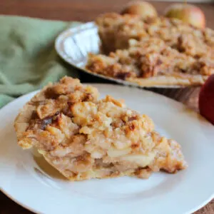 Slice of apple crumble pie with bottom crust, apple slices and butter crumb topping on top served on small white plate.