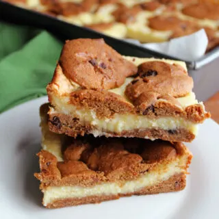 Two pieces of chocolate chip cookie cheesecake bars stacked on top of each other showing cookie crust, creamy cheesecake filling and more cookies on top.