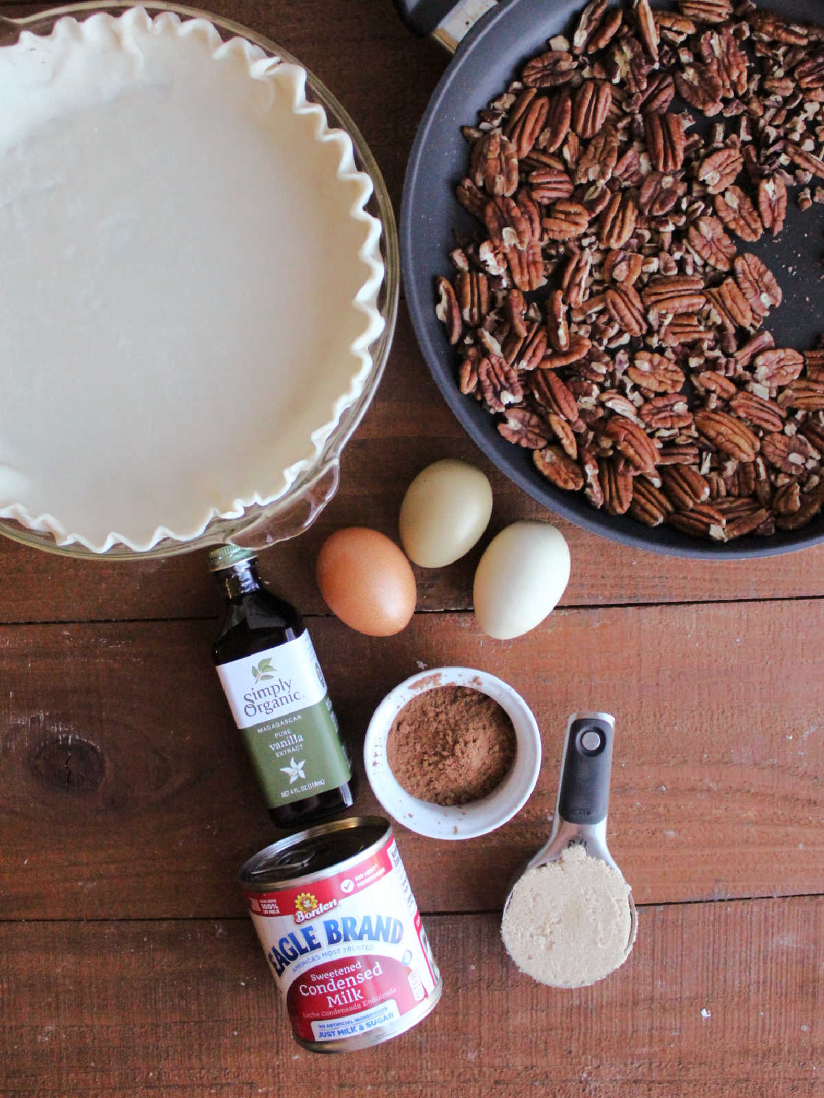 Ingredients including toasted pecans, pie crust, sweetened condensed milk, vanilla, cocoa powder, brown sugar and eggs, ready to be made into chocolate pecan pie.