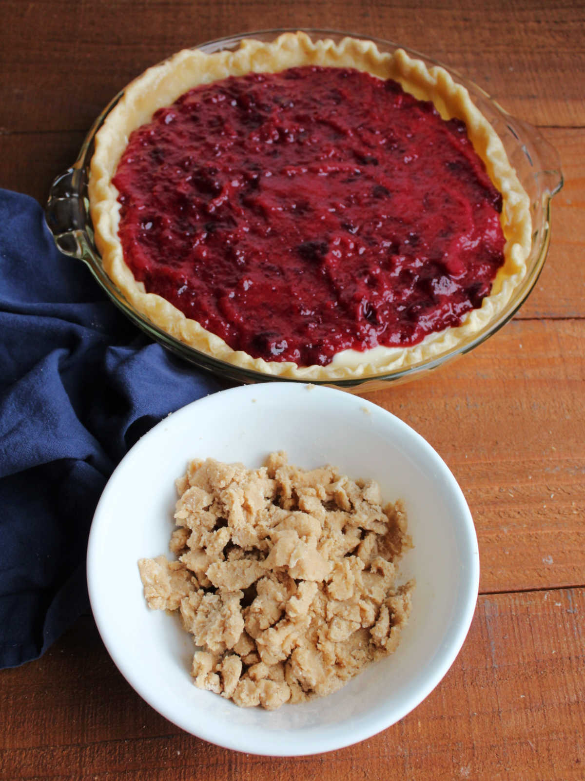 Small bowl filled with brown sugar crumb mixture in front of pie pan with crust, cream cheese mixture, and cranberry mixture inside. 