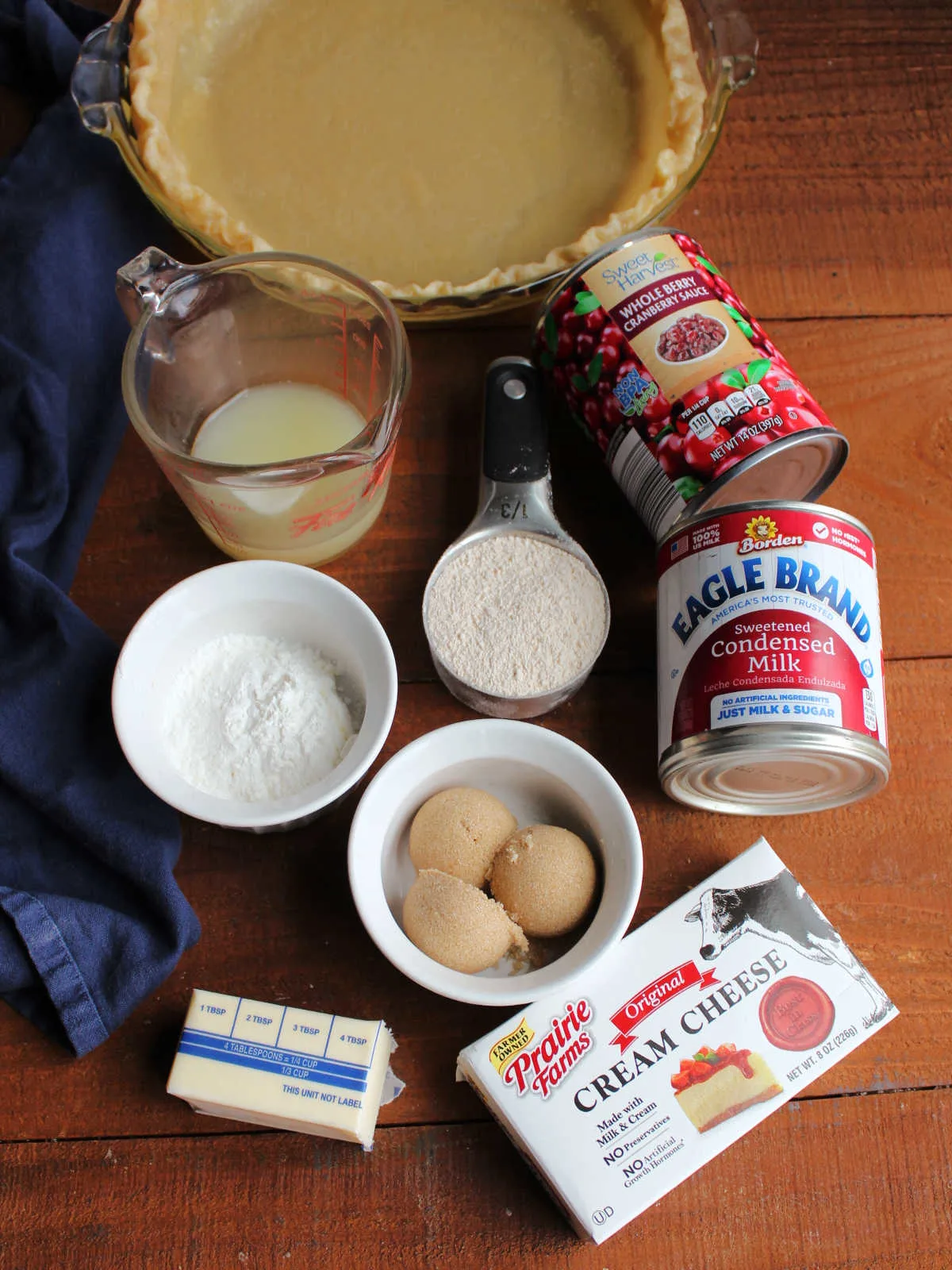 Ingredients including partially baked pie crust, cranberry sauce, sweetened condensed milk, cream cheese, brown sugar, flour, cornstarch, butter, and lemon juice ready to be made into a cranberry crumble pie.
