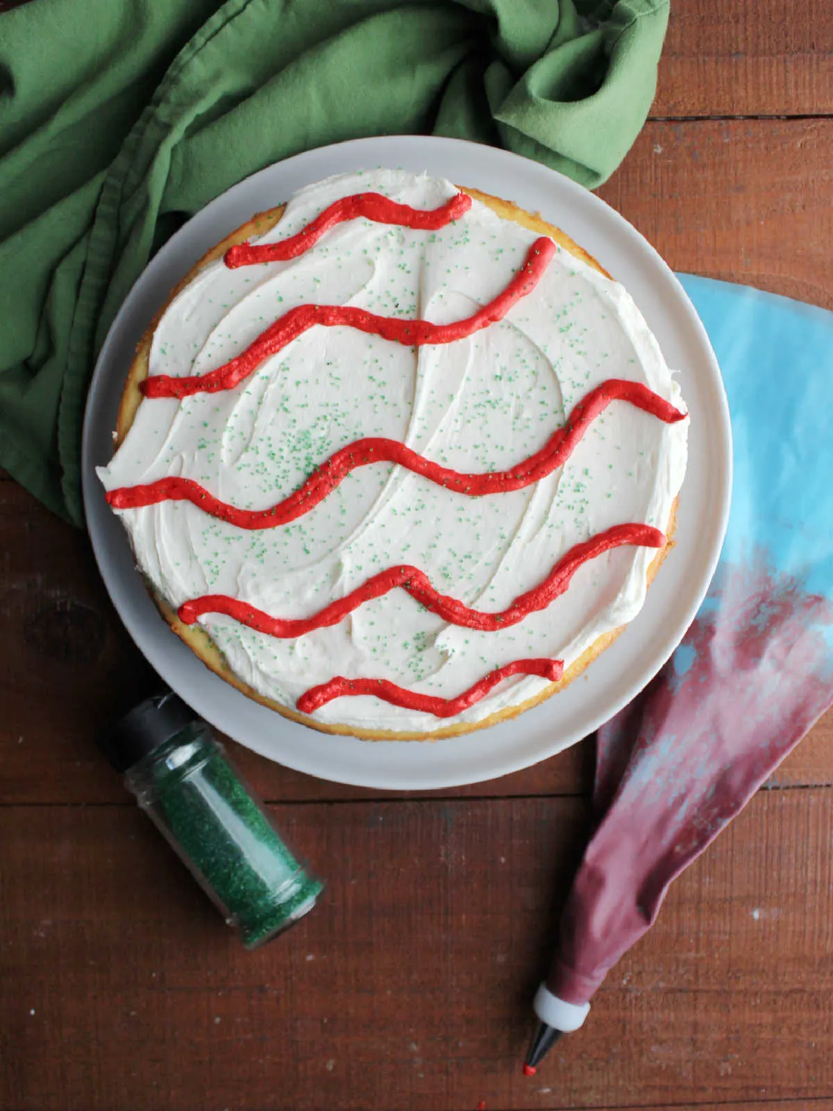 Decorated cheesecake next to piping bag with red frosting and bottle of green sanding sugar.