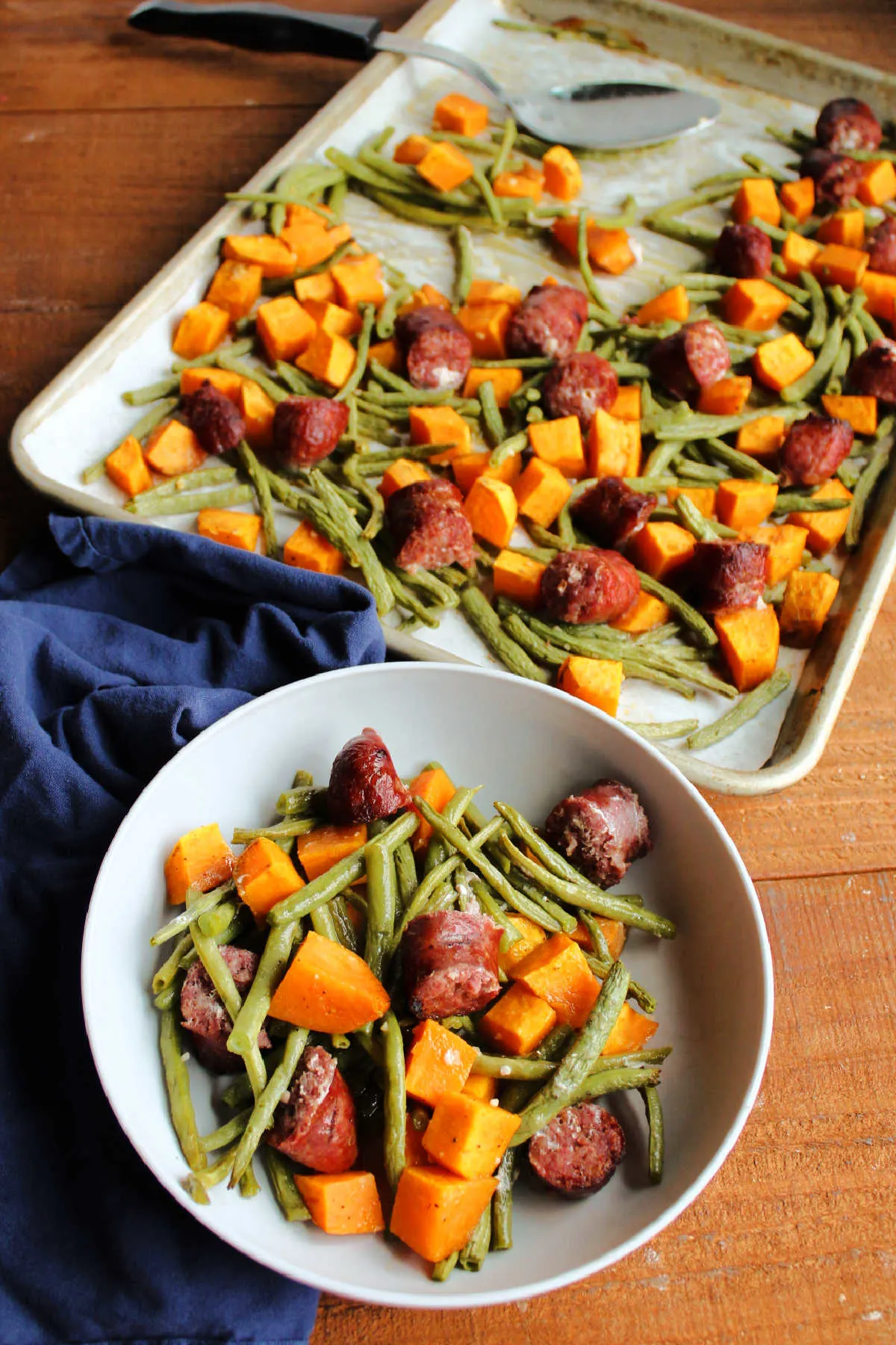 Bowl of roasted sweet potatoes, green beans and sausage ready to eat with sheet pan of remaining food nearby.
