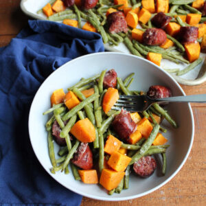 Bowl of roasted green beans, sweet potatoes, and smoked sausage with fork ready to eat and sheet pan with remaining food in background.