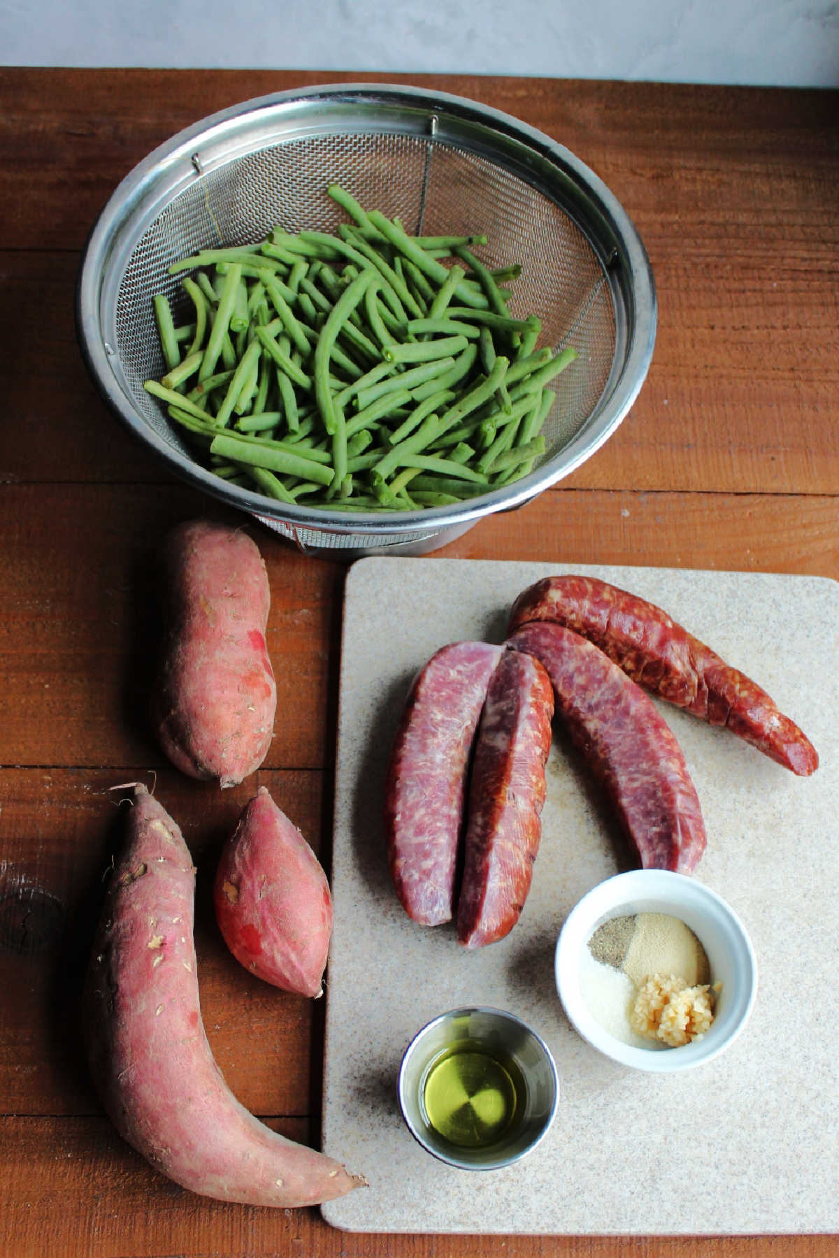 Ingredients including smoked sausage, fresh sweet potatoes and green beans, oil, garlic, salt, pepper, and onion powder ready to be made into sheet pan supper.
