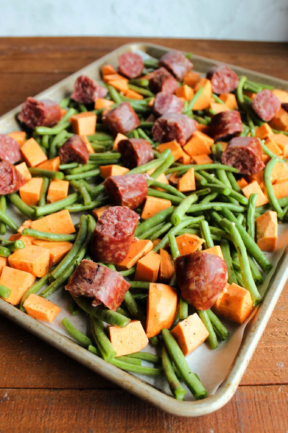 Chunks of sausage placed over green beans and sweet potatoes on sheet pan, ready to go in the oven. 