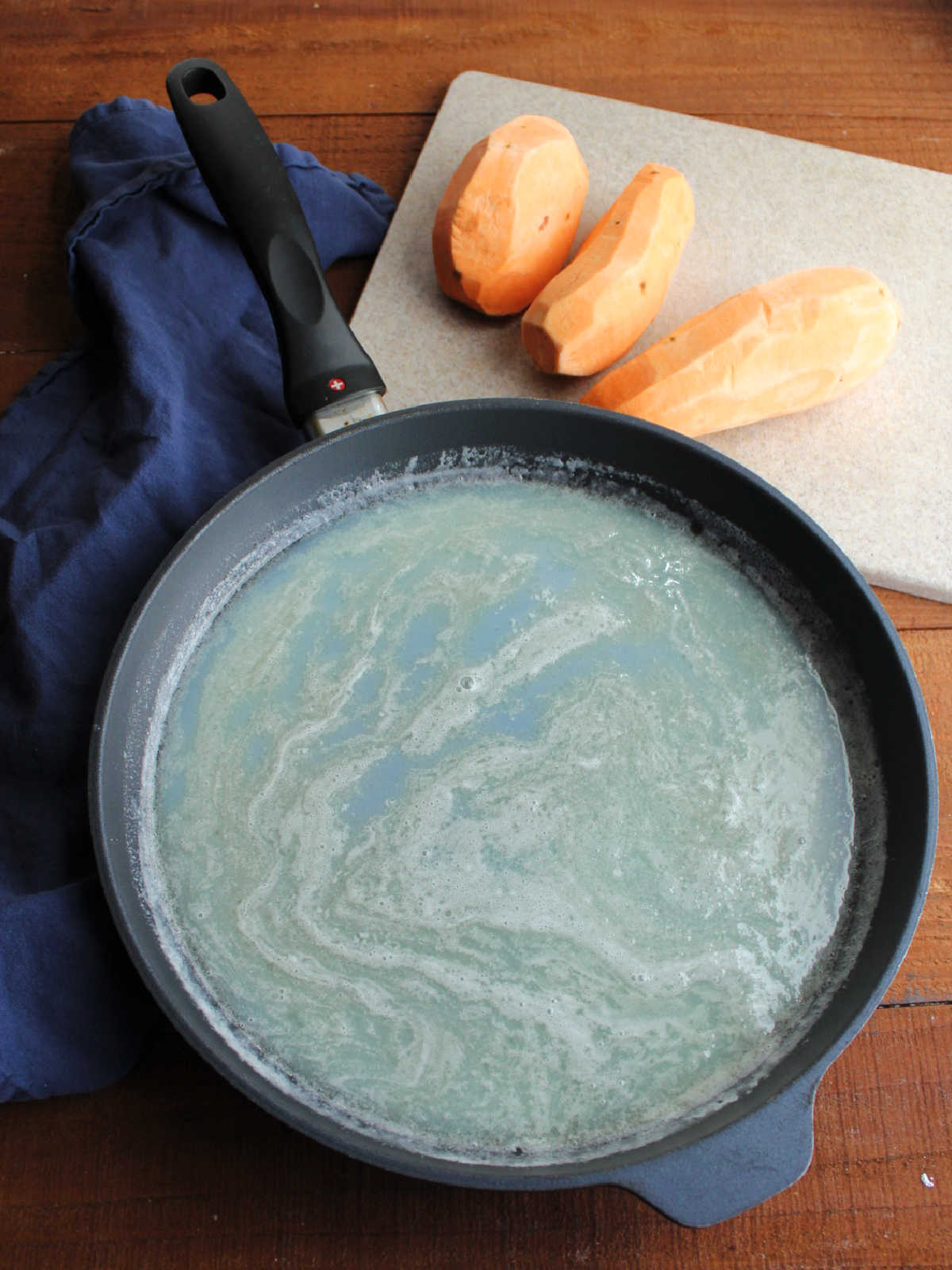 Large skillet with melted butter, sugar, and milk mixture next to peeled sweet potatoes on a cutting board.