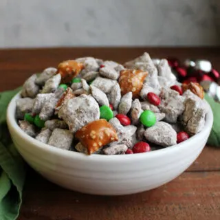 White serving bowl filled with Christmas Chex mix with chocolate and peanut butter muddy buddies, peanut butter filled pretzels, powdered sugar and holiday M&Ms.