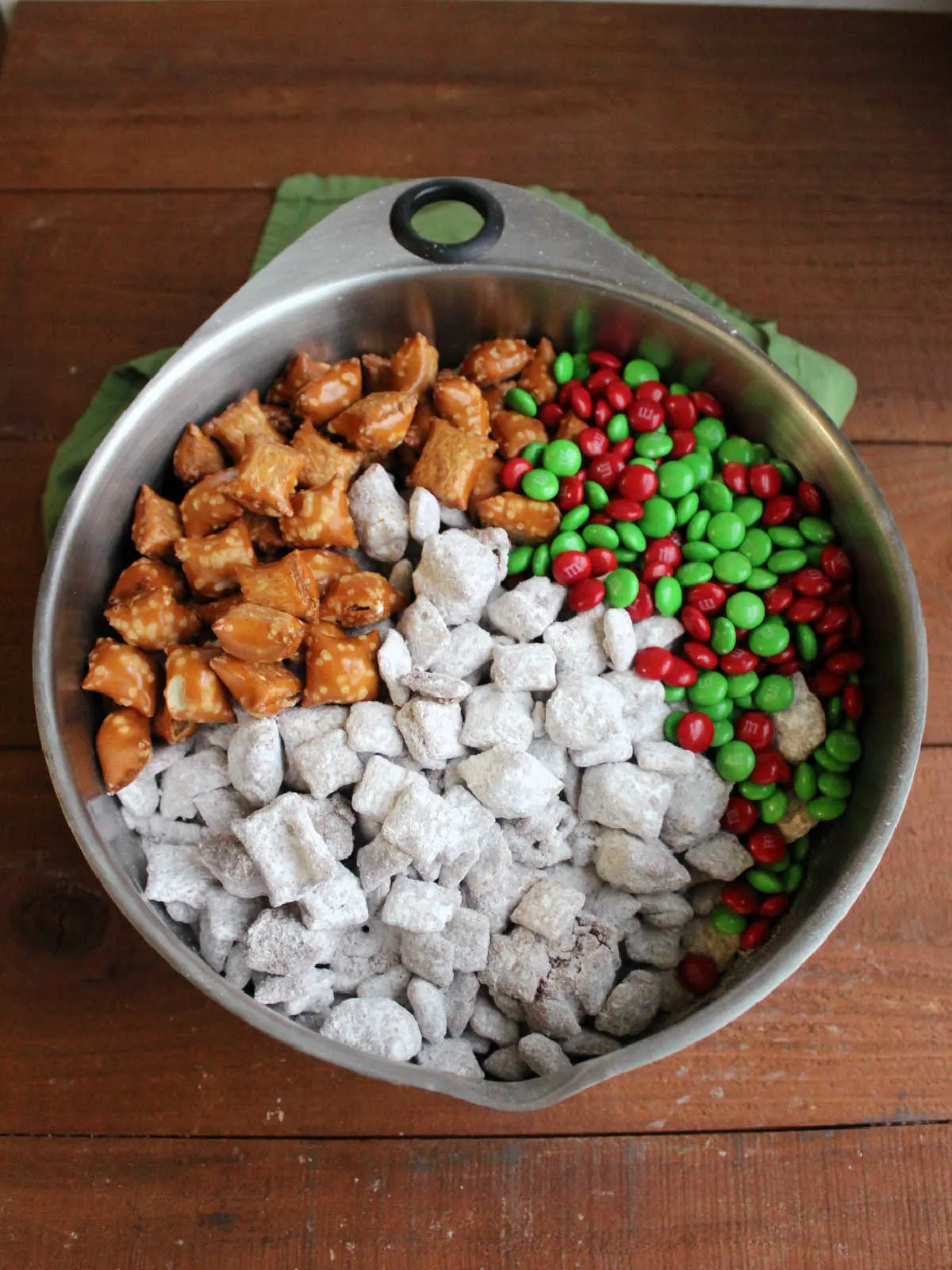 Powdered sugar coated puppy chow in large bowl with holiday M&Ms and more peanut butter filled pretzels. 