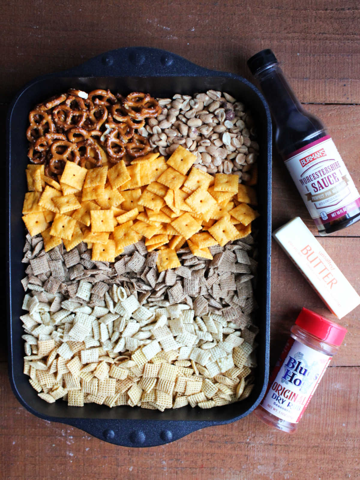 Roasting pan with 3 varieties of Chex cereal, cheese crackers, peanut, and pretzels inside next to Worcestershire sauce, butter, and Blues Hog bbq rub showing ingredients needed to make this snack mix.