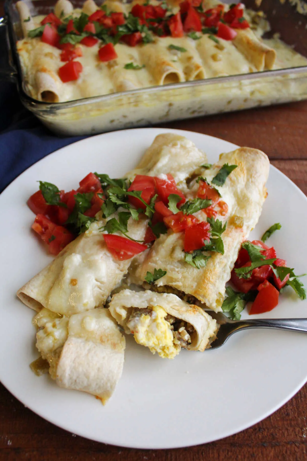 Bite of breakfast enchilada on fork showing scrambled eggs and sausage inside flour tortilla and white cheese sauce on top.