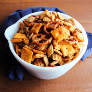 Antique white bowl filled with smokey bbq chex mix made from cheese crackers, cereal, peanuts, and pretzels.