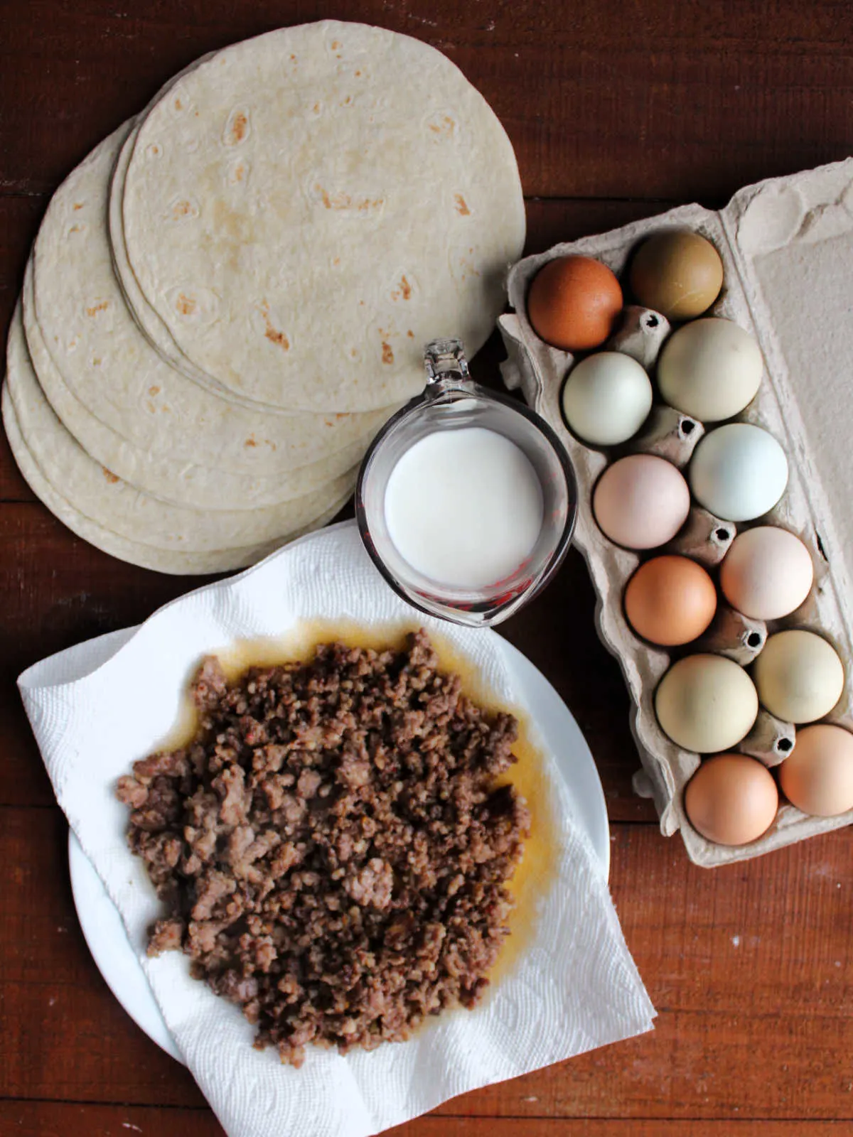 Ingredients including a dozen eggs, milk, browned sausage, and tortillas ready to be made into breakfast enchiladas.