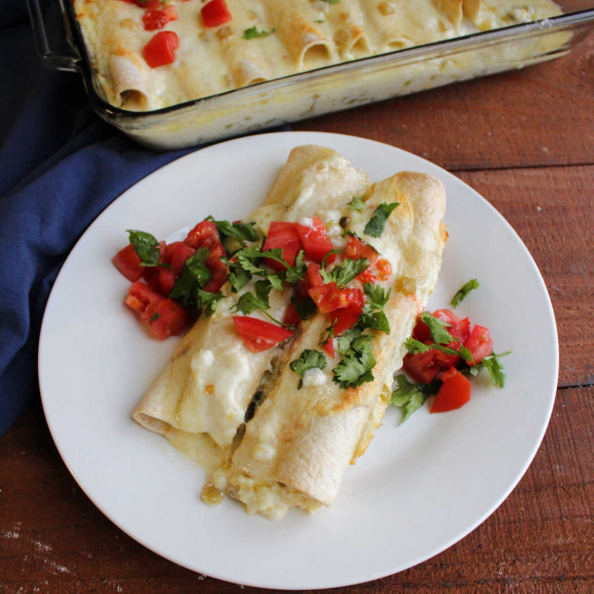 Plate with two breakfast enchiladas baked in creamy cheese sauce and topped with diced tomato and fresh cilantro, ready to eat. 