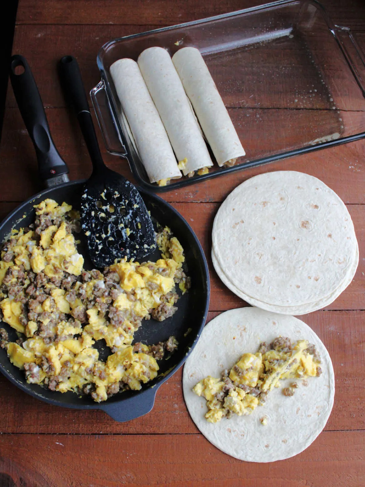 Skillet with sausage and egg mixture next to pile of tortillas, one with some sausage and eggs on it and some enchiladas in a baking dish nearby showing how to fill the tortillas and roll them up.