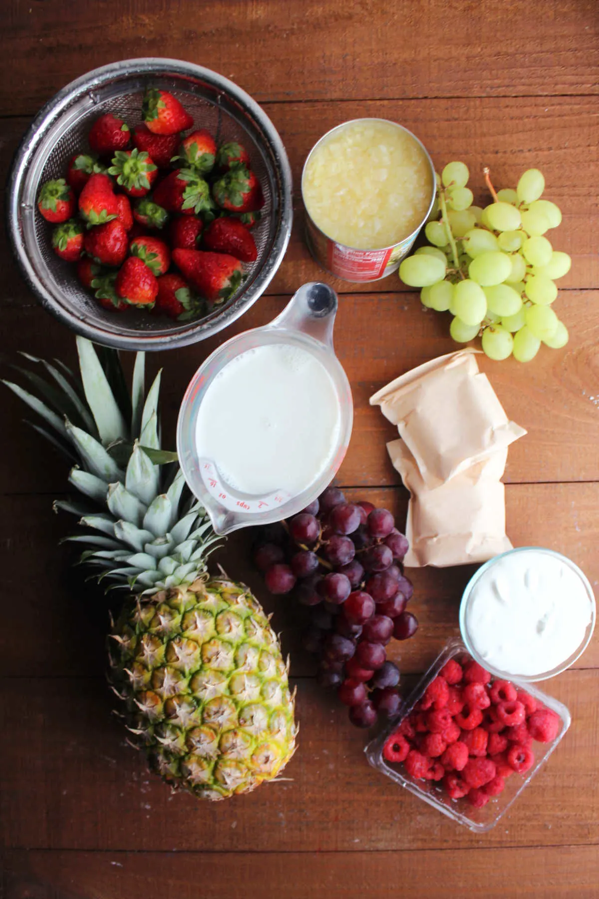 Ingredients including pineapple, strawberries, crushed pineapple, grapes, instant pudding mix, milk, sour cream, and raspberries, ready to be made into fruit salad.