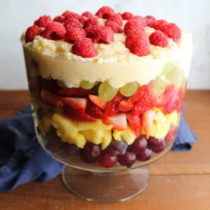Glass trifle bowl filled with layered fruit salad with red grapes, pineapple, strawberries, green grapes, pudding mixture, and raspberries on top.