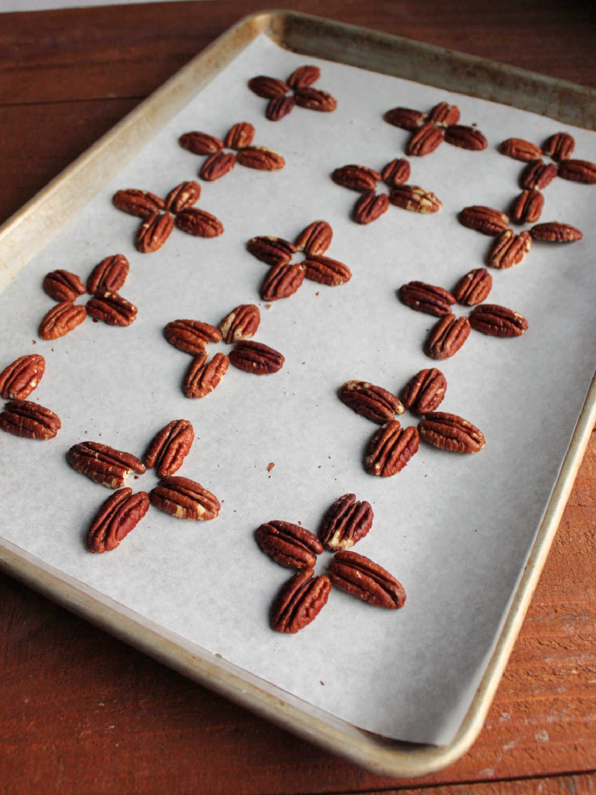 Parchment paper lined baking sheet with groups of 4 toasted pecans in cross formations to be made into turtles.