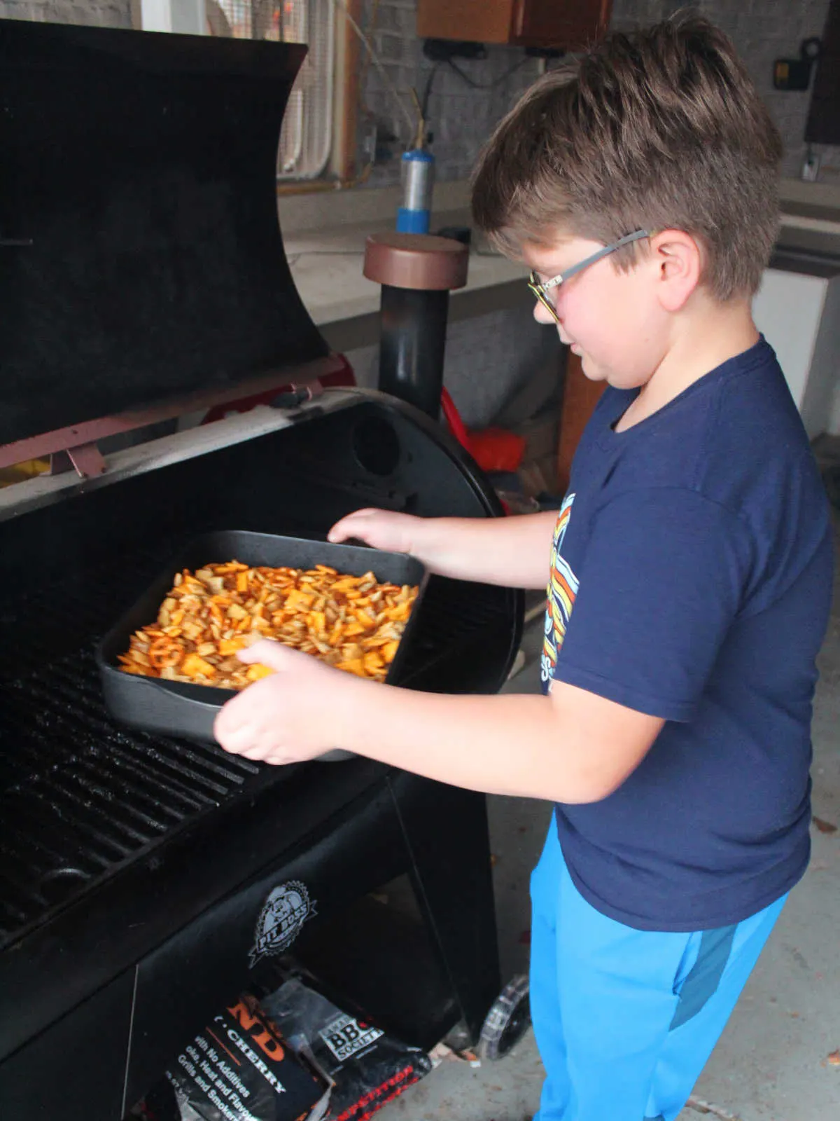 Child putting roasting pan of Chex mix mixture onto a pellet grill style smoker.