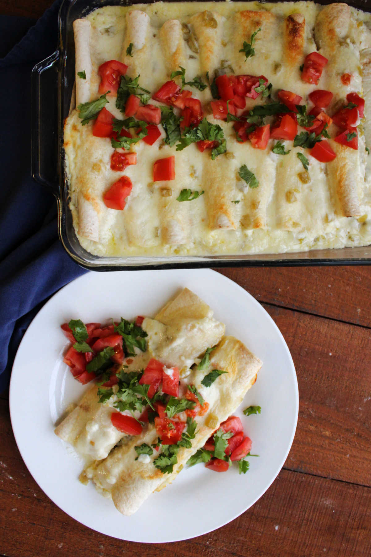Pan of breakfast enchiladas next to a plate with two enchiladas topped with extra tomato and cilantro, ready to eat. 
