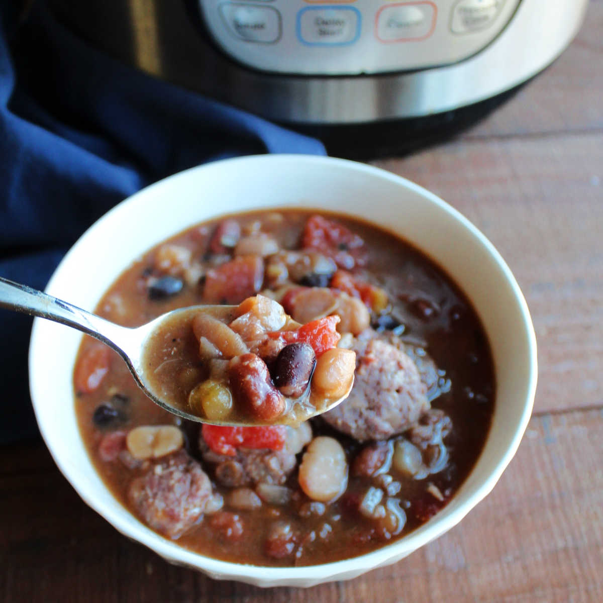 Spoonful of bean soup with tomatoes and kielbasa over bowl of soup next to Instant Pot. 