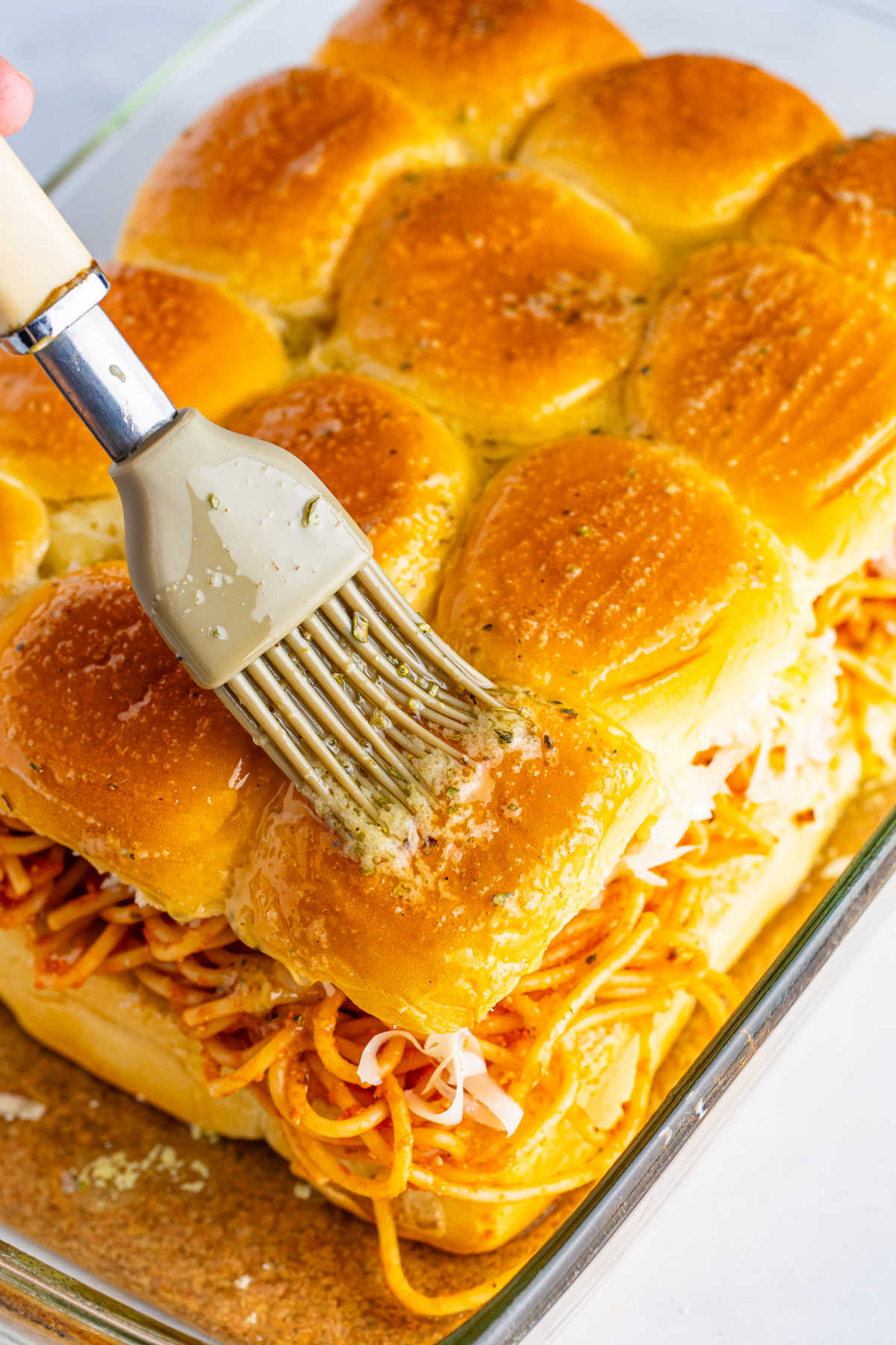 Brushing garlic butter over the top of the slider buns with spaghetti and cheese showing in the middle of the sliders.
