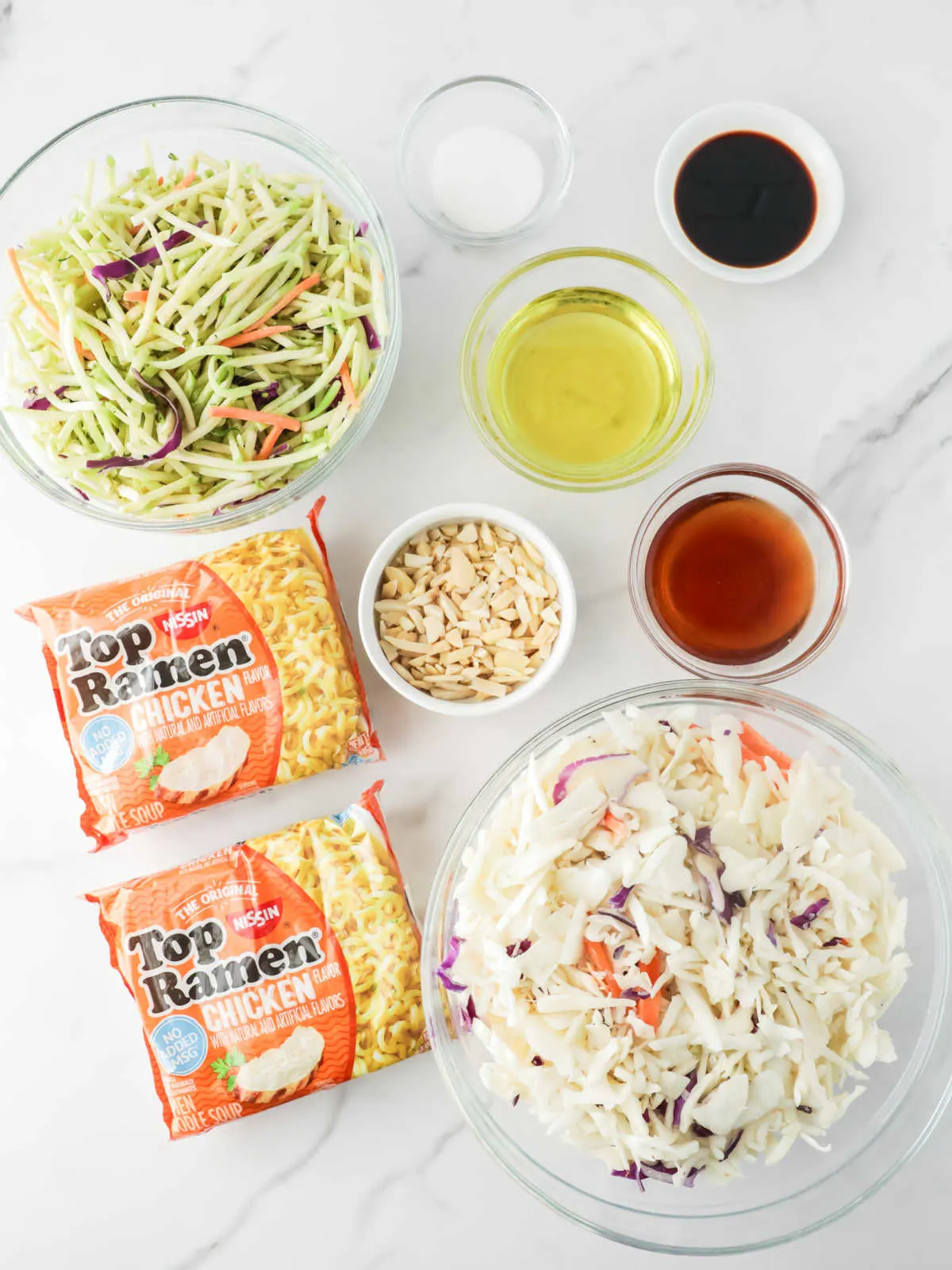 Ingredients including broccoli slaw mix, coleslaw mix, ramen noodles, slivered almonds, soy sauce, olive oil, sugar, and rice vinegar ready to be made into ramen slaw.