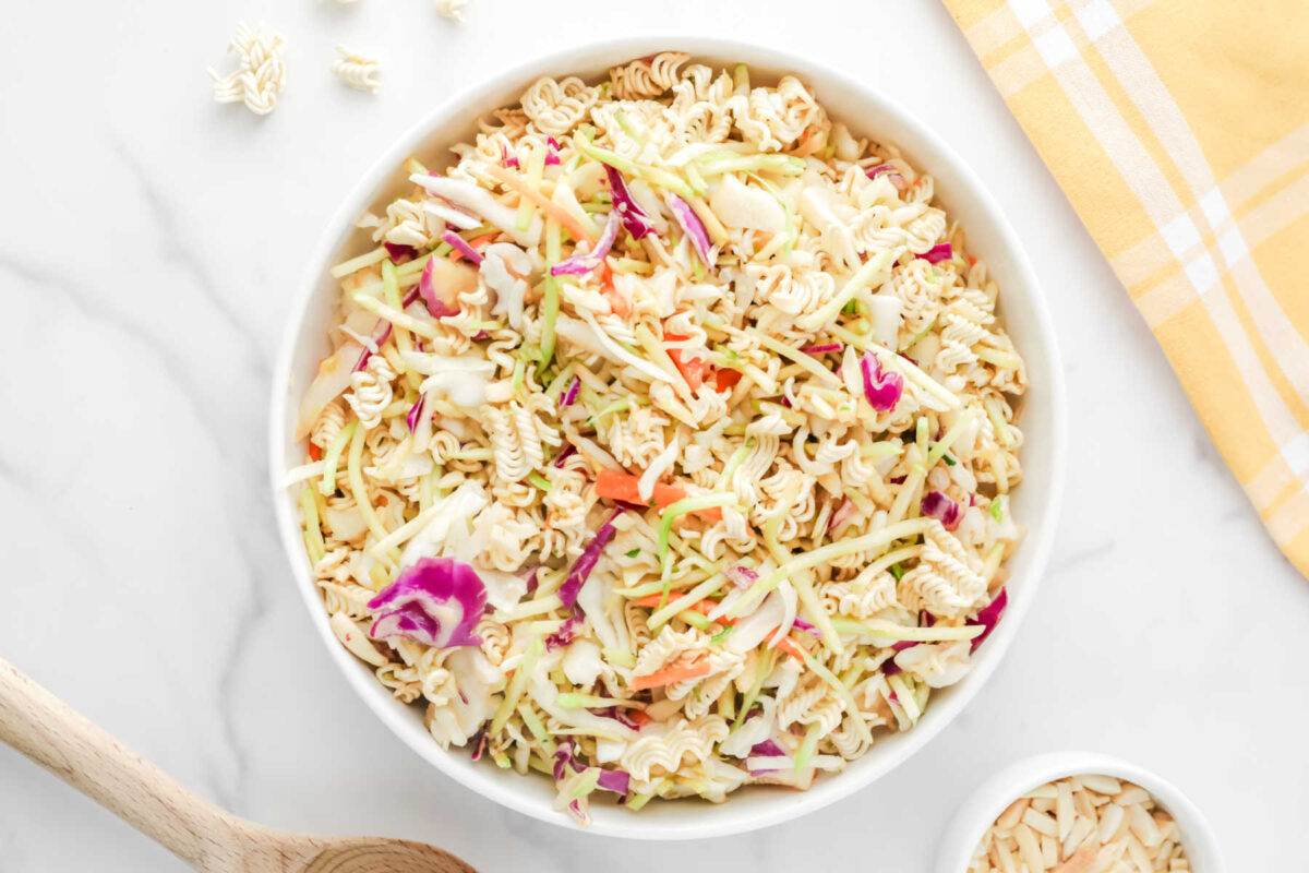 Large serving bowl filled with coleslaw mix of cabbage and broccoli with bits of crunchy ram noodles and slivers of almonds in a light dressing.