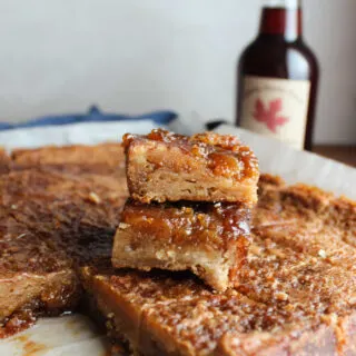 Maple syrup bars with golden brown sugar crust and gooey maple syrup filling stacked on top of each other with bottle of pure maple syrup in the background.